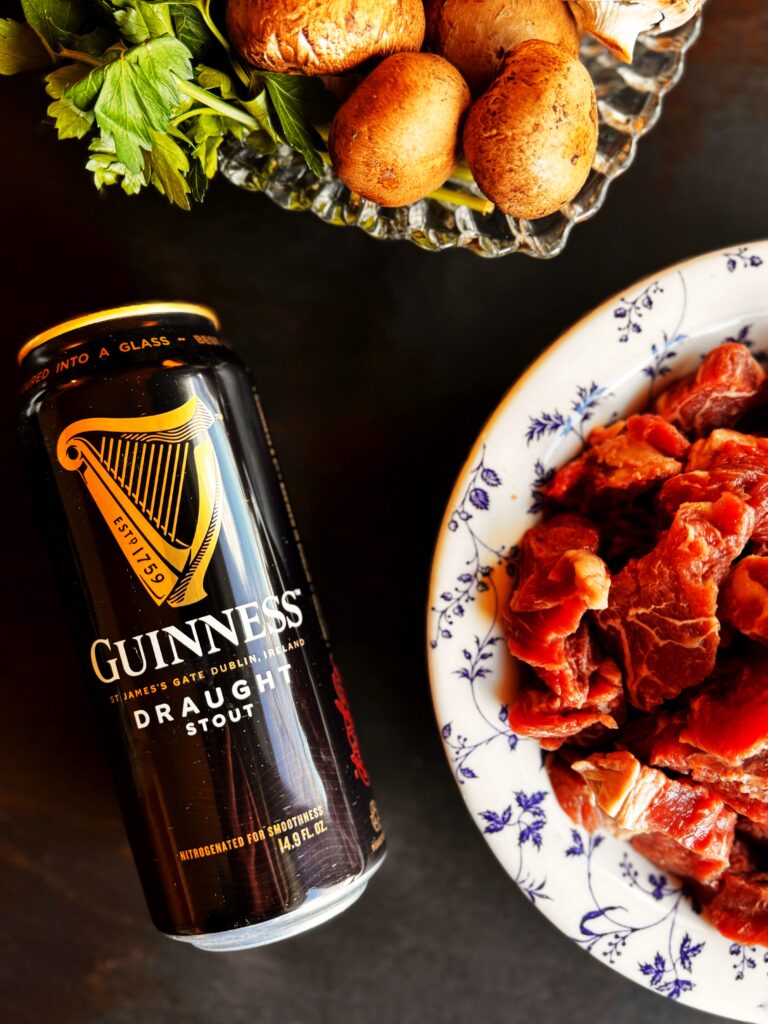 photo of a can of Guinness beer, a bowl of stew meat, and a bowl of portobello mushrooms and fresh parsley for beef Guinness Stew