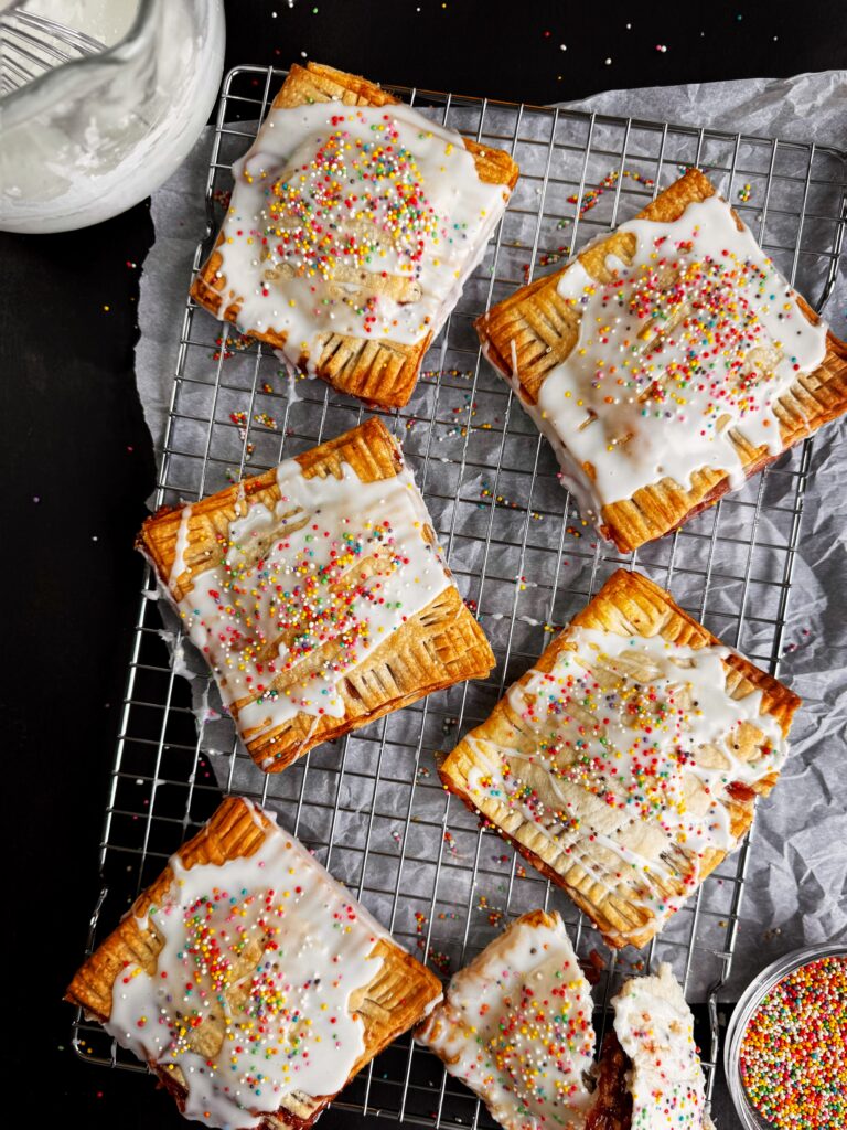 photo of homemade strawberry almond pop tarts sitting on a wire rack and parchment paper with bowl of white icing and a bowl of rainbow sprinkles