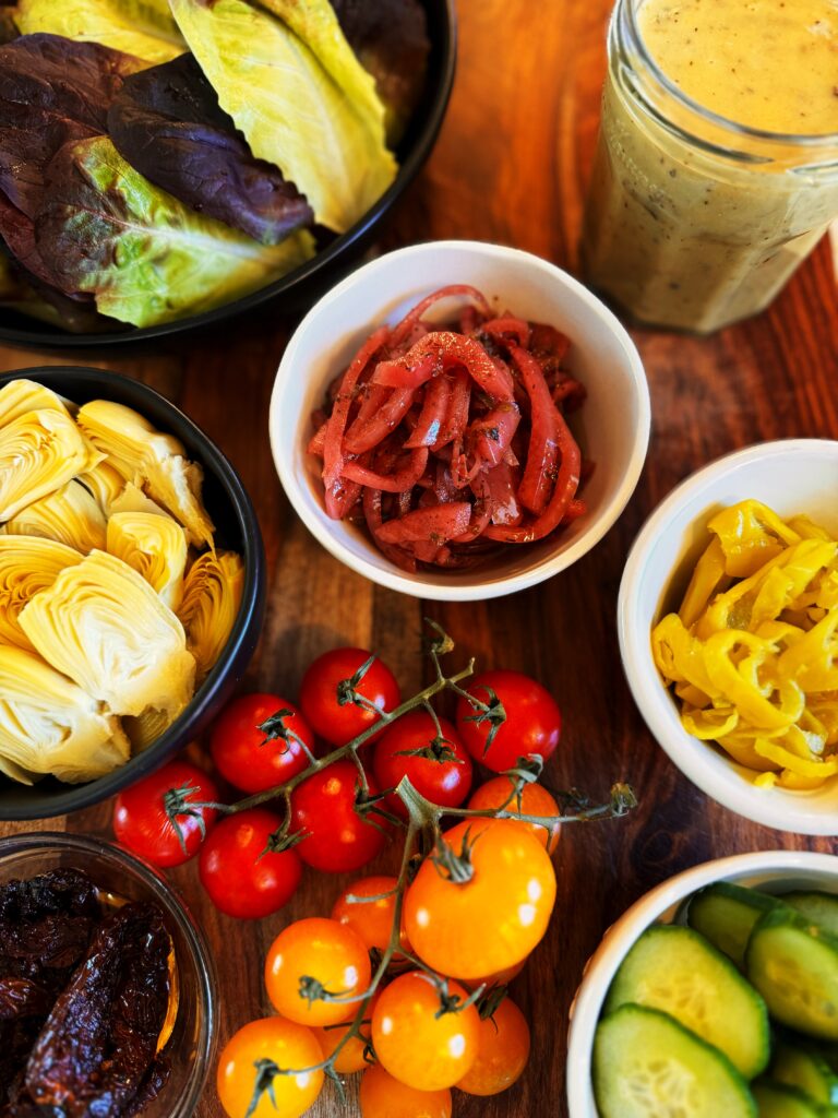 photo of red and yellow cherry tomatoes on the vine, a bowl of baby leaf lettuce, a jar of lemon tahini salad dressing, and small bowls of artichoke hearts, sliced pepperoncini, and sliced cucumbers