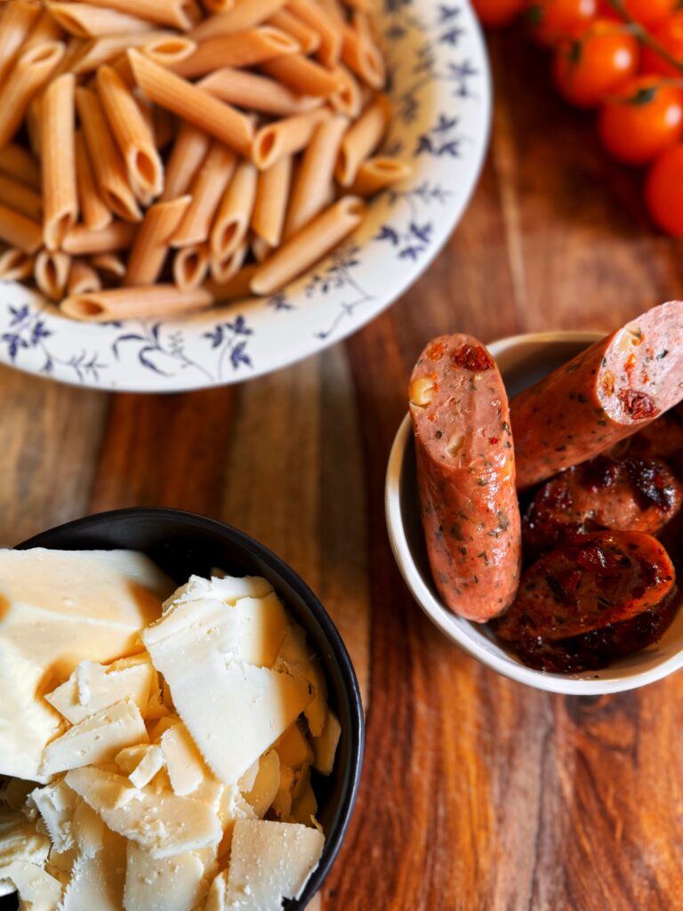 photo of bowls of shaved asiago cheese, sliced chicken sausage, and cooked whole wheat penne pasta