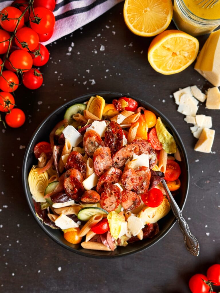 a photo of a large bowl of green salad with pasta, chicken sausage, and lemon tahini dressing. The bowl is surrounded by a display of red cherry tomatoes on the vine, half of a lemon, shaved Asiago cheese, and flaked sea salt