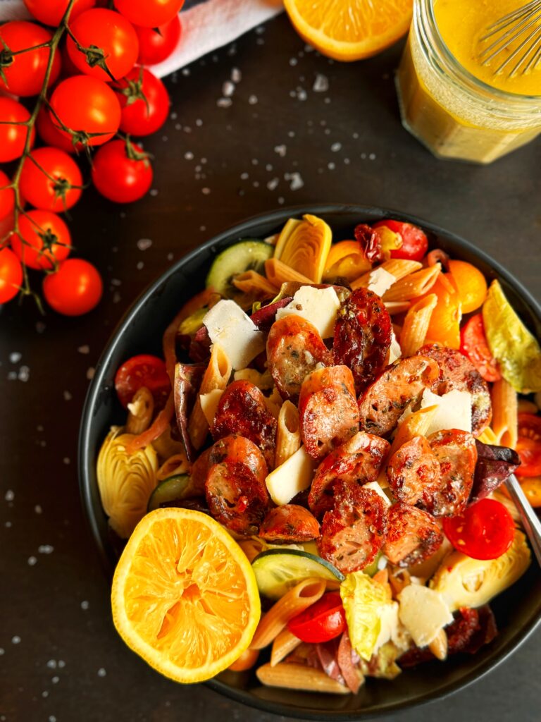 a photo of a large bowl of green salad with pasta, chicken sausage, and lemon tahini dressing. The bowl is surrounded by a display of red cherry tomatoes on the vine, half of a lemon, shaved Asiago cheese, and flaked sea salt