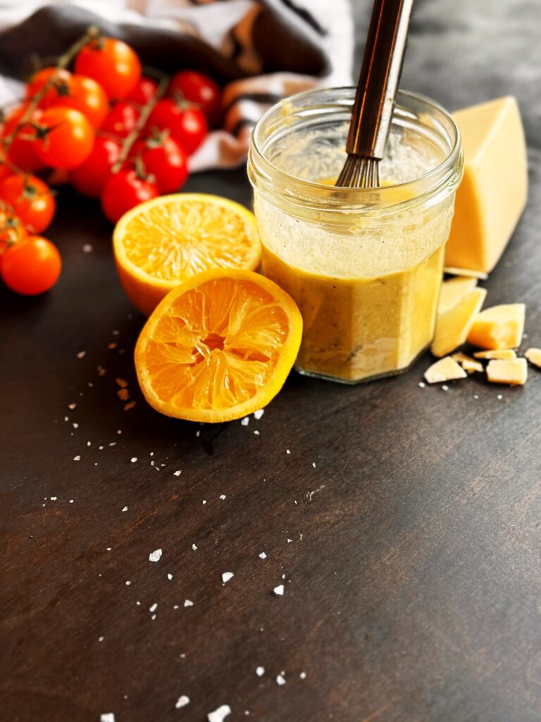 photo of a glass jar of lemon tahini salad dressing and two lemon halves, red cherry tomatoes on the vine, and a wedge of Asiago cheese