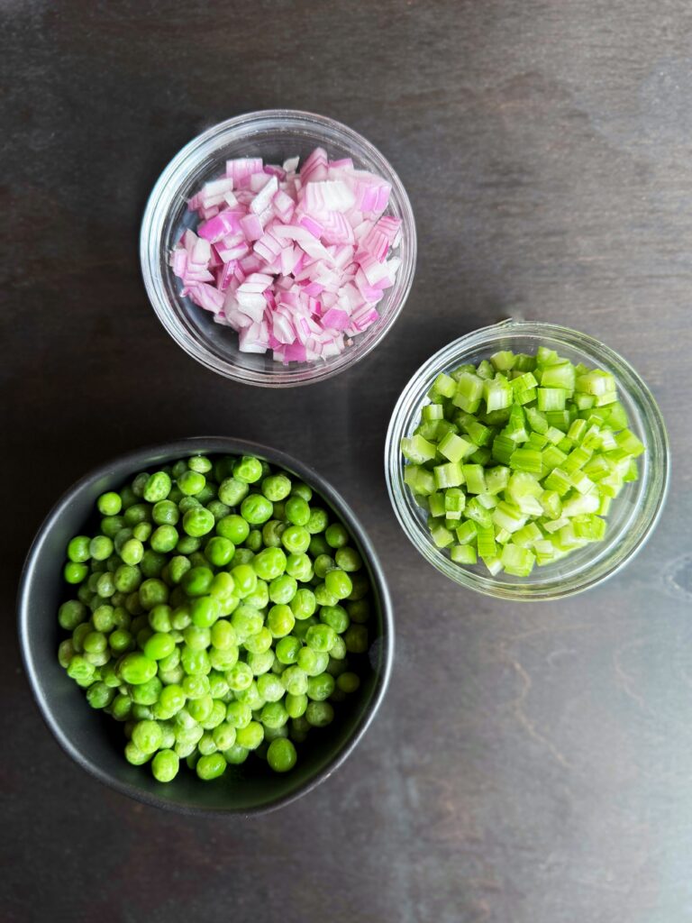 a photo of one small black and two small clear bowls sitting on a dark brown tabletop. The clear bowls are filled with diced red onion and diced celery and the black bowl is filled with green peas