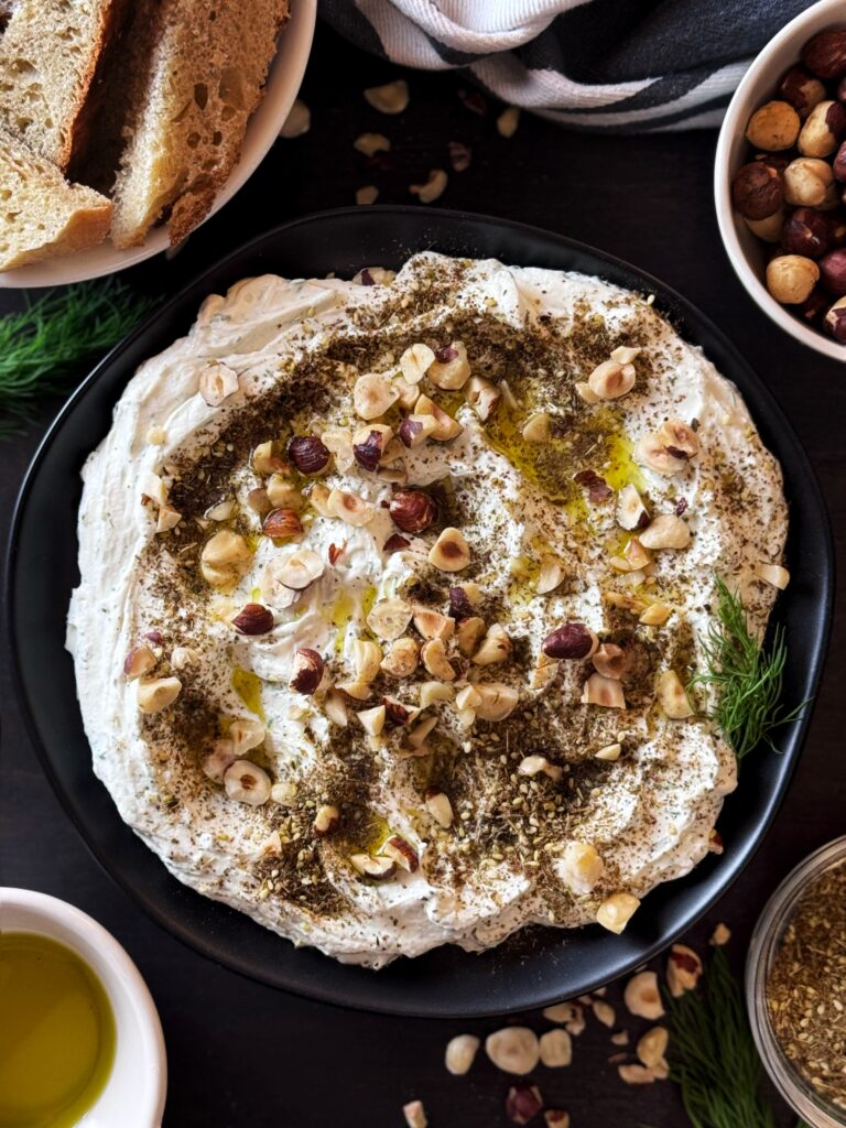 photo of whipped goat cheese spread on a black ceramic plate and topped with a drizzle of olive oil and a sprinkling of chopped hazelnuts and za'atar seasoning