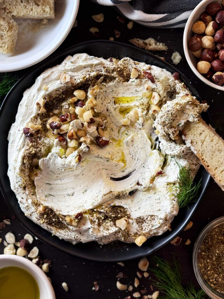 photo of whipped goat cheese spread on a black ceramic plate and topped with a drizzle of olive oil and a sprinkling of chopped hazelnuts and za'atar seasoning. There is also a piece of bread dipped into the whipped goat cheese.