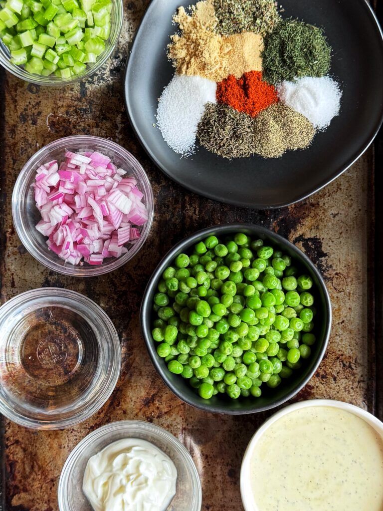 photo of bowls filled with ingredients for classic creamy pasta salad with peas. The bowls are black, white, and clear, and are setting on top of a rusty cooking sheet. The bowls are filled with green peas, diced red onion, diced celery, herbs and spices, mayonnaise, vinegar, and yoghurt