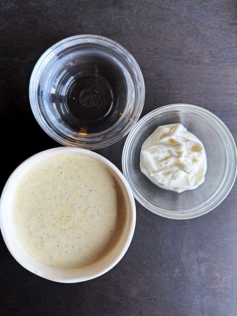 a photo of two small clear bowls and one small white bowl sitting on top of a dark brown tabletop. The clear bowls are filled with white vinegar and yoghurt. The white bowl is filled with mayonnaise.