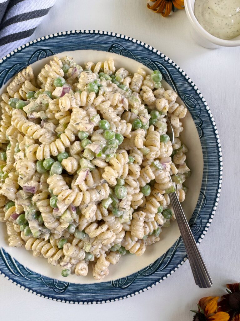 a photo of a large blue and white bowl that is filled with creamy pasta salad with peas. The bowl is sitting on a white tabletop an a silver spoon is stuck in the bowl of pasta salad