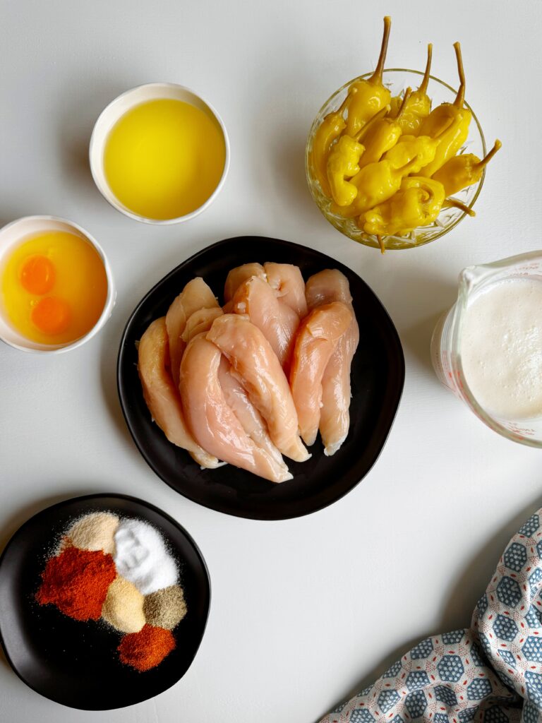 photo of a plate of raw chicken breast tenders and bowls sitting on a white tabletop and filled with pepperoncini peppers, raw eggs, buttermilk, and spices