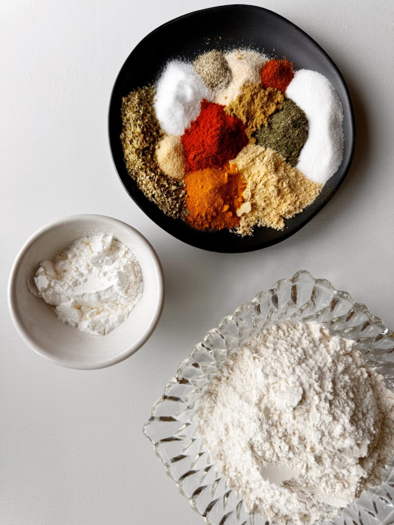 photo of a black plate filled with different spices, a clear crystal bowl filled with flour, and a small white bowl filled with cornstarch, all sitting on a white tabletop