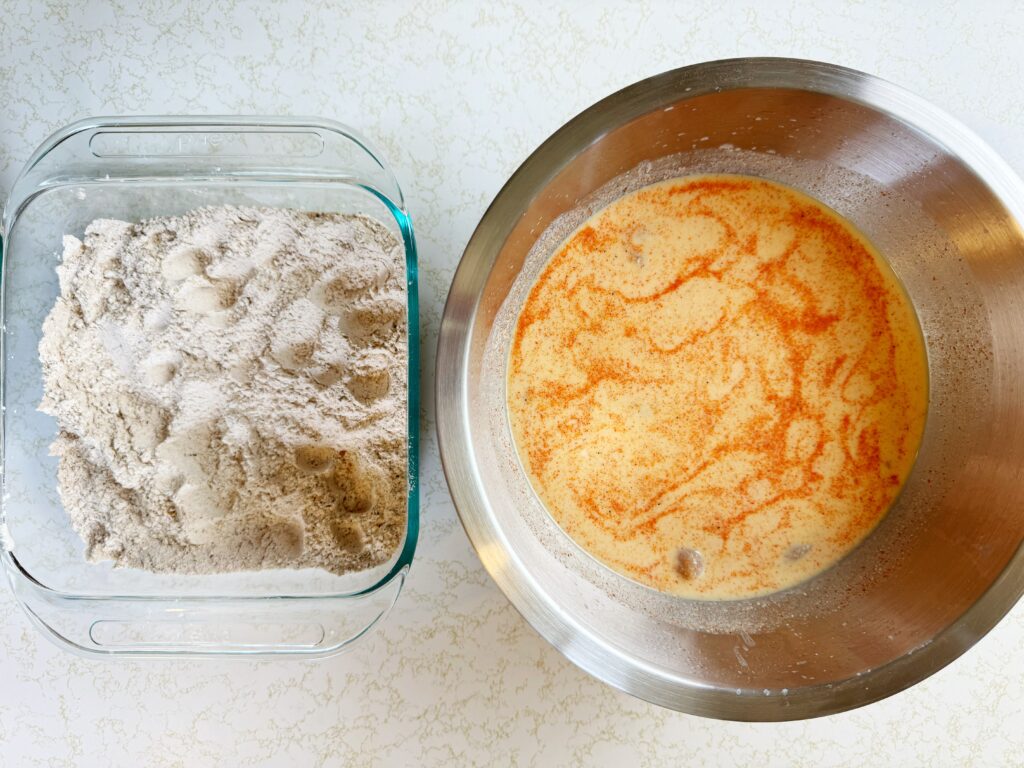 a photo of a large metal bowl full of raw chicken tenders in a buttermilk brine and a glass baking dish full of all-purpose flour