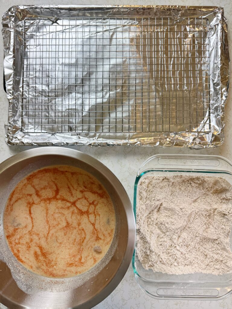 a photo of a large metal bowl full of raw chicken tenders in a buttermilk brine, a glass baking dish full of all-purpose flour, and a wire rack sitting on an aluminum foil-lined baking sheet